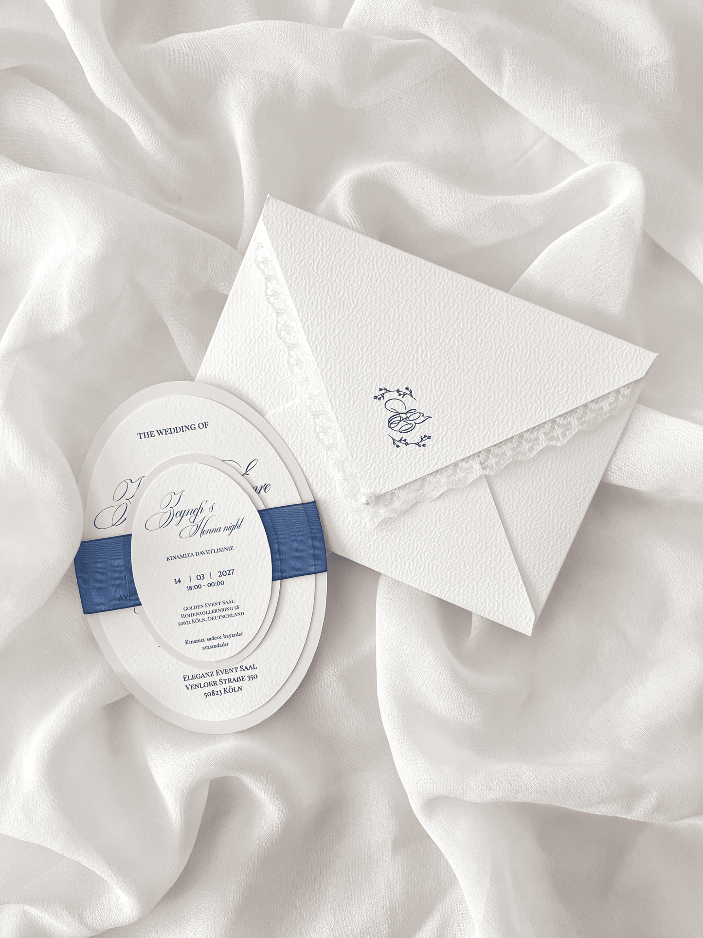 Faire-part de mariage personnalisé Collection MONARCH avec enveloppe blanche à contour dentelle, liner intérieur avec monogramme, invitation ovale doublée, carte RSVP assortie et large ruban bleu foncé.