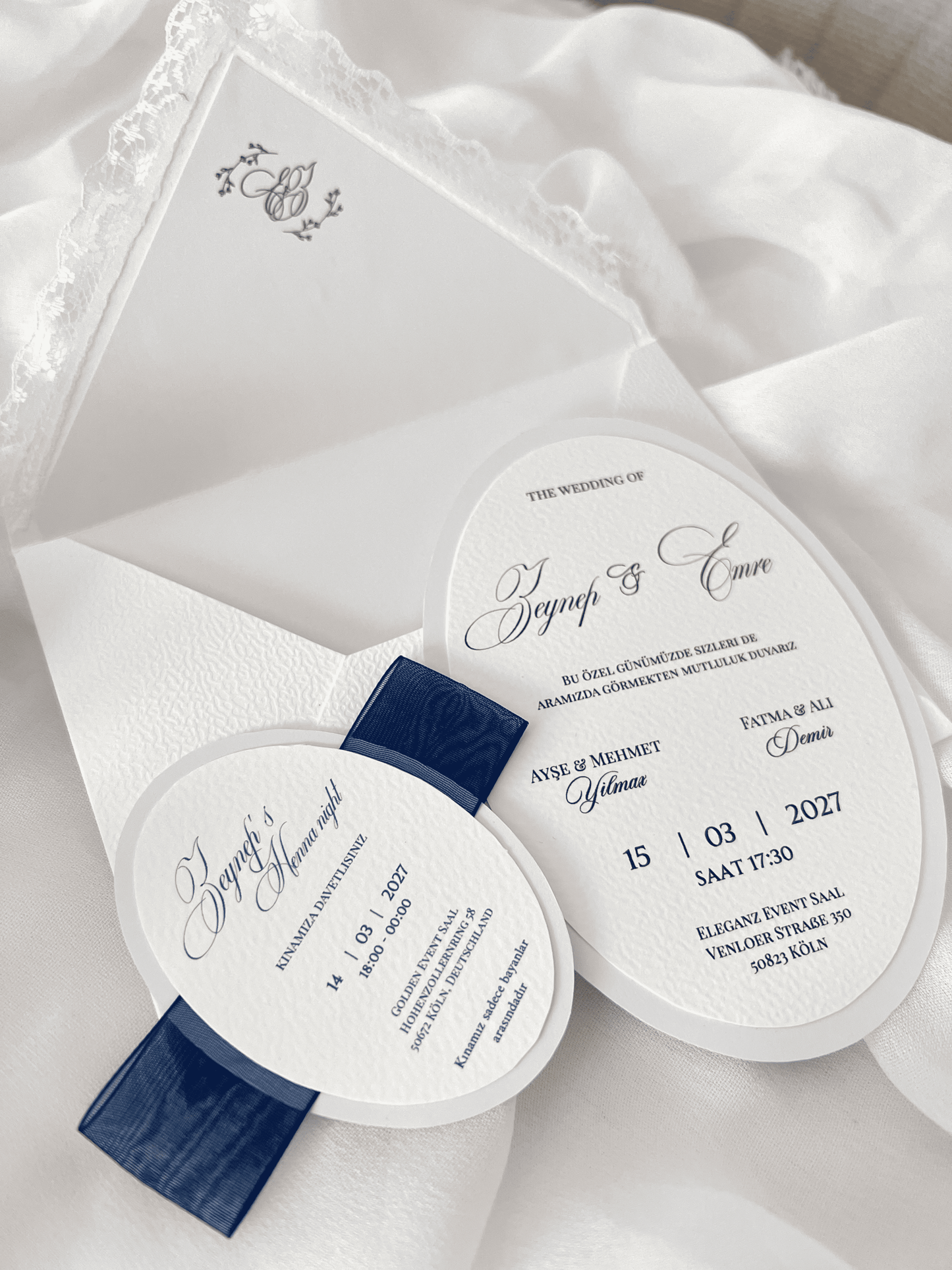 Faire-part de mariage personnalisé Collection MONARCH avec enveloppe blanche à contour dentelle, liner intérieur avec monogramme, invitation ovale doublée, carte RSVP assortie et large ruban bleu foncé.