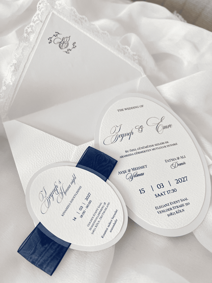 Faire-part de mariage personnalisé Collection MONARCH avec enveloppe blanche à contour dentelle, liner intérieur avec monogramme, invitation ovale doublée, carte RSVP assortie et large ruban bleu foncé.