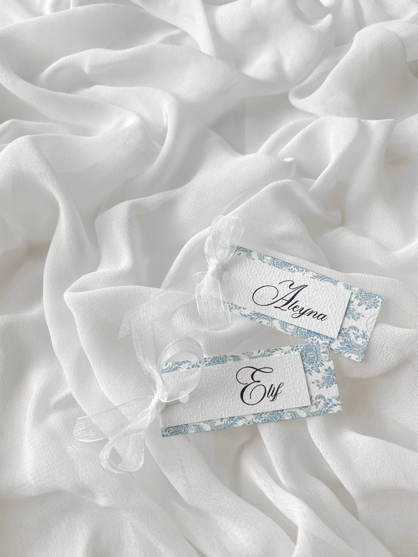Marque-place de mariage personnalisé Collection WISTERIA I composé d'une carte florale bleu pastel superposée à une carte blanche avec prénom personnalisé, disponible avec ou sans ruban ou gland.