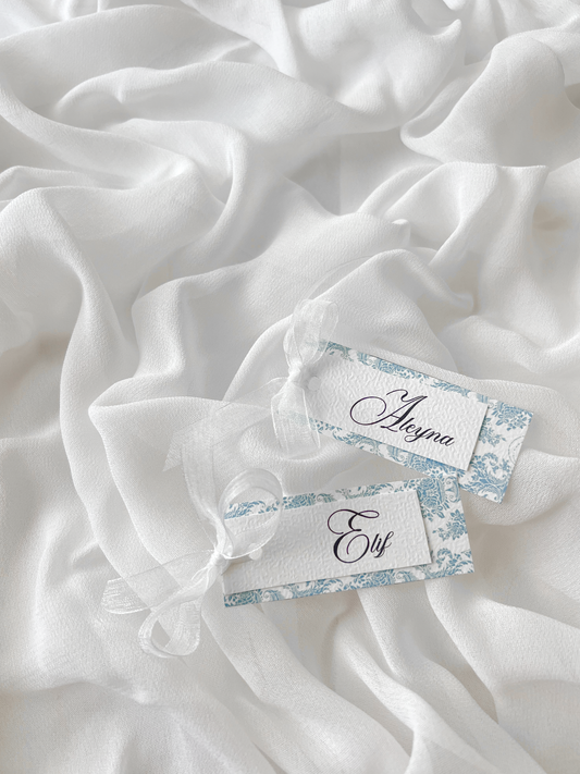 Marque-place de mariage personnalisé Collection WISTERIA I composé d'une carte florale bleu pastel superposée à une carte blanche avec prénom personnalisé, disponible avec ou sans ruban ou gland.